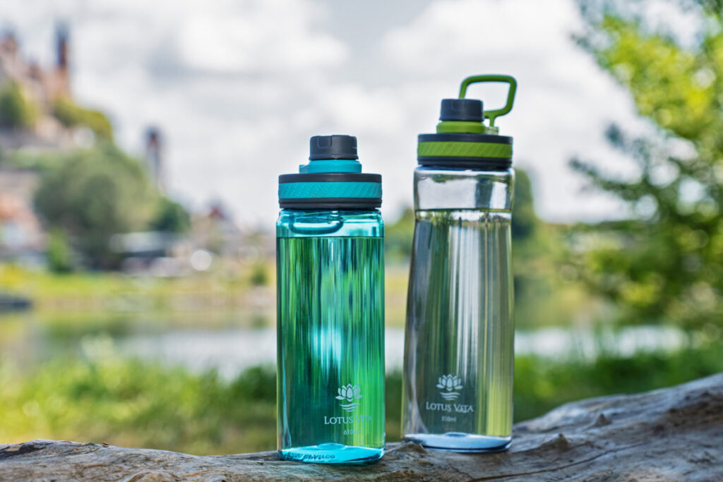 Zwei Lotus Vita Trinkflaschen, eine blaugrün und eine durchsichtig, stehen auf einem Baumstamm vor einer unscharfen Flusslandschaft.