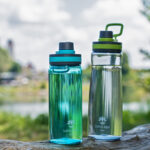 Zwei Lotus Vita Trinkflaschen, eine blaugrün und eine durchsichtig, stehen auf einem Baumstamm vor einer unscharfen Flusslandschaft.