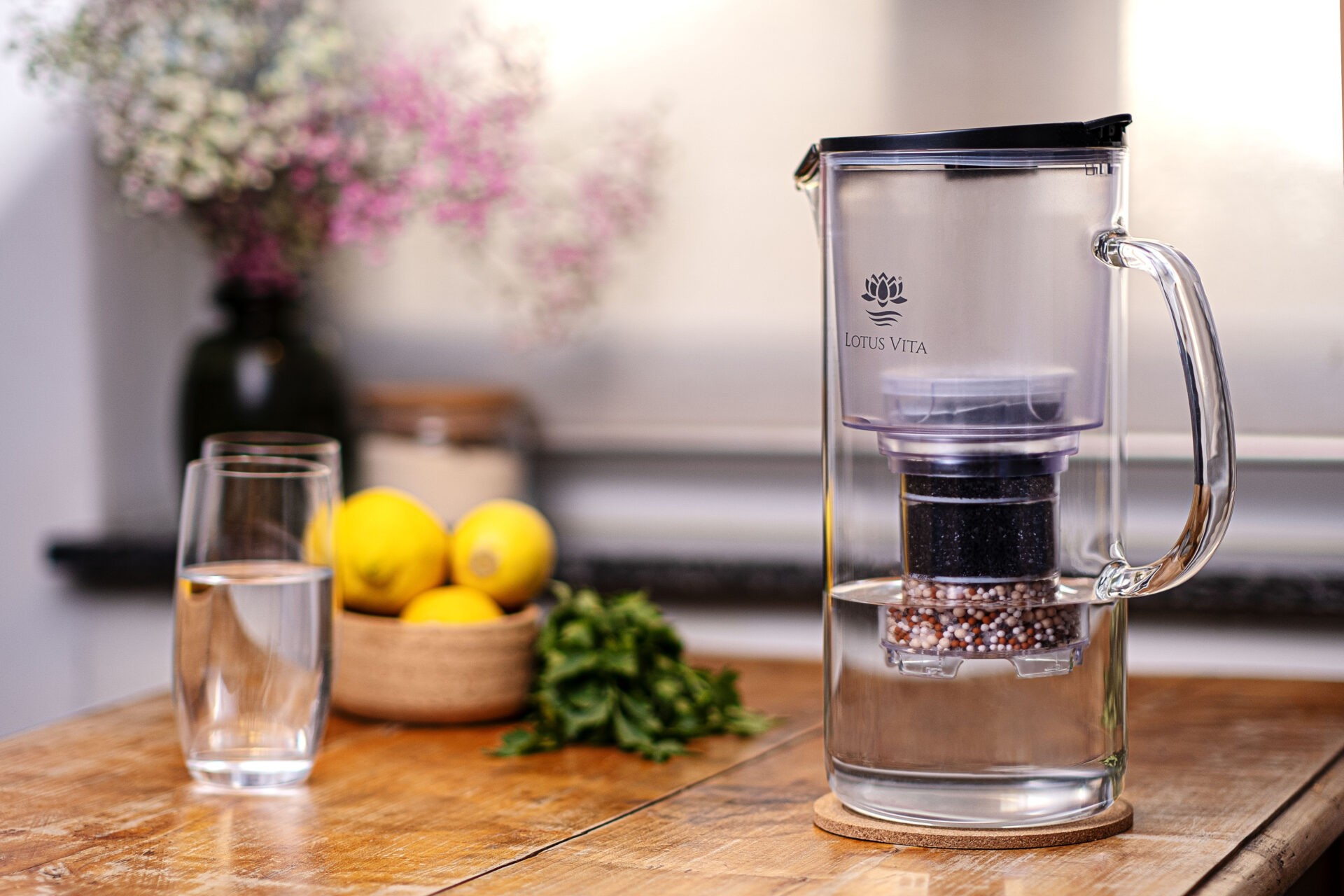 Eine gläserne Wasserfilterkanne mit Filterkartusche steht auf einem Holztisch neben einem gefüllten Wasserglas und frischen Zitronen.
