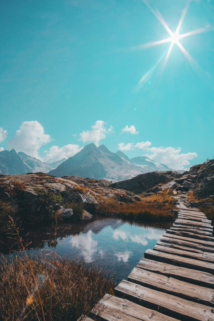 Ein Holzsteg führt über einen klaren Bergsee, in dem sich der sonnige Himmel spiegelt, vor einer beeindruckenden Bergkulisse.