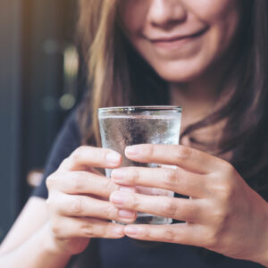 Eine lächelnde Frau hält ein Glas mit frischem, kaltem Wasser in beiden Händen und genießt den Moment der Erfrischung.