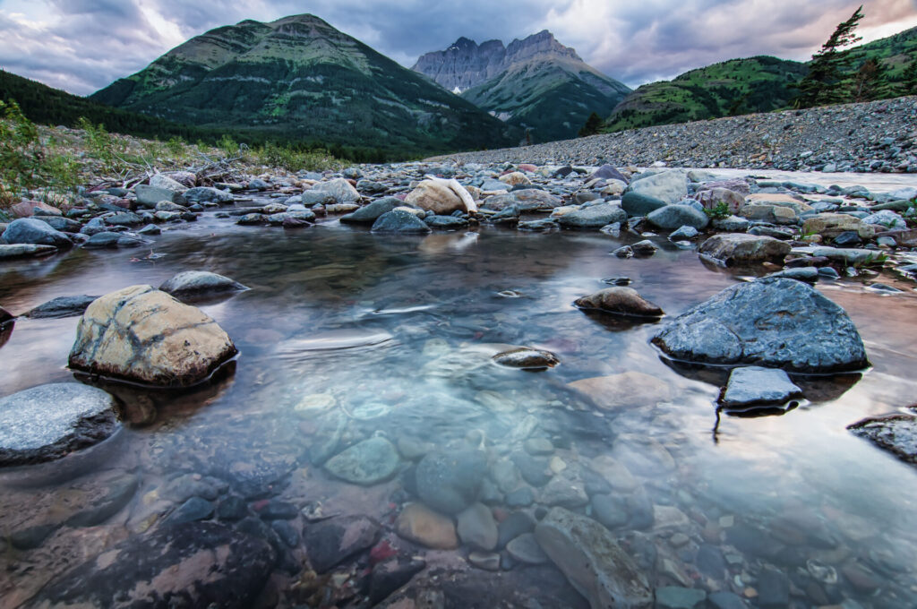 Klares Wasser eines Bergflusses fließt über Kieselsteine, im Hintergrund eine beeindruckende Bergkette.