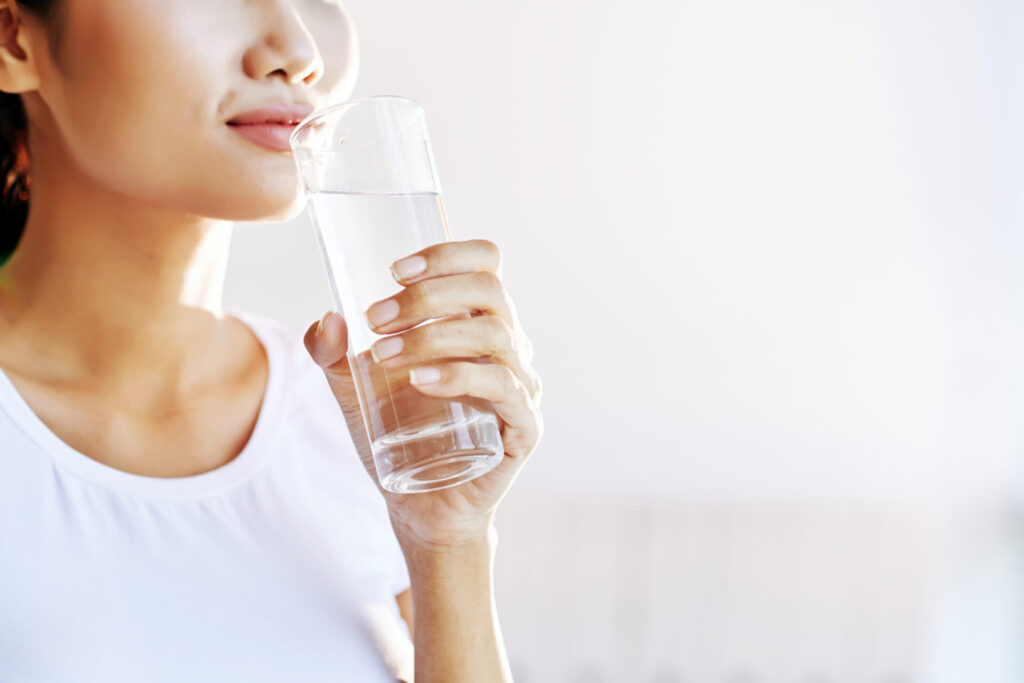 Eine Frau in einem weißen T-Shirt führt ein Glas mit klarem Wasser zum Mund, um zu trinken.