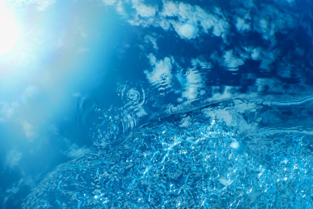 Klares, blaues Wasser, in dem sich die Sonne und weiße Wolken am Himmel spiegeln und leichte Wellen schlagen.