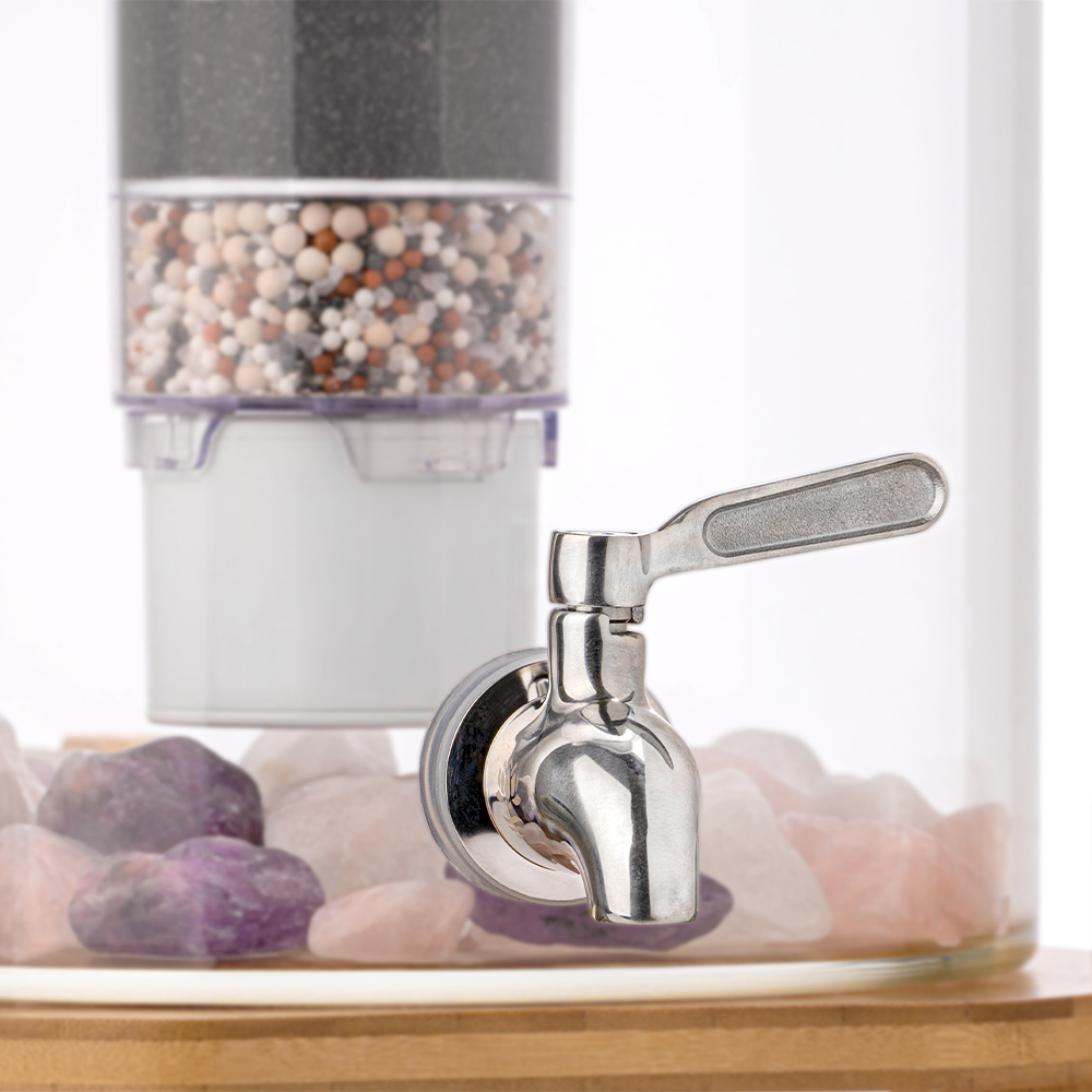 Nahaufnahme eines Edelstahl-Wasserhahns an einem Glas-Wasserspender, im Hintergrund eine mehrstufige Filterkartusche und am Boden Edelsteine.