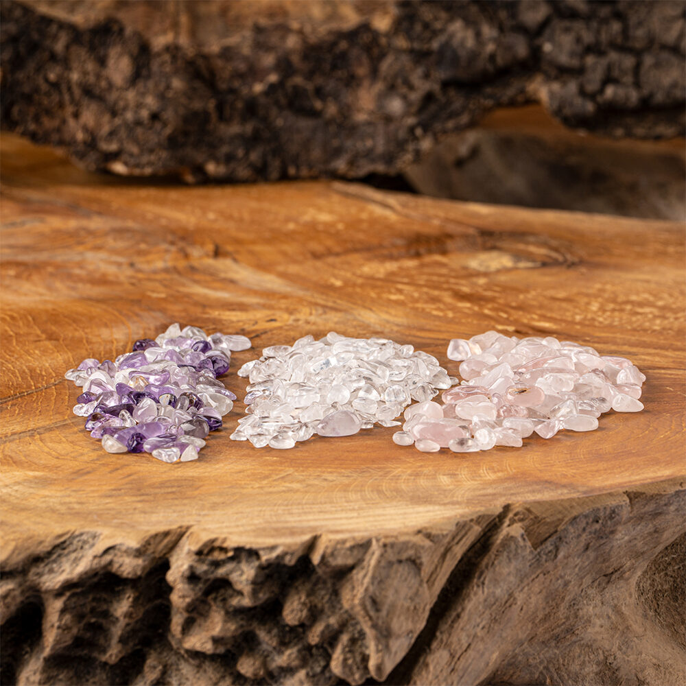 Drei kleine Haufen der Edelsteine Rosenquarz, Bergkristall und Amethyst liegen auf einer rustikalen Holzoberfläche.