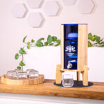 Ein Glas-Wasserfilter mit Bambusgehäuse und -ständer steht auf einer Holztheke, daneben ein Tablett mit Gläsern und eine grüne Pflanze im Hintergrund.