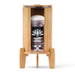 Ein Glas-Wasserfilter mit mehrstufiger Filterkartusche und Edelsteinen, eingefasst in einen Ständer mit Lichtschutz und Deckel aus hellem Bambus.