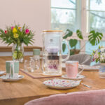 Eine Wasserfilter-Karaffe aus Glas steht im Mittelpunkt eines festlich gedeckten Holztisches, umgeben von Tassen und bunten Blumensträußen in einer wohnlichen Atmosphäre.