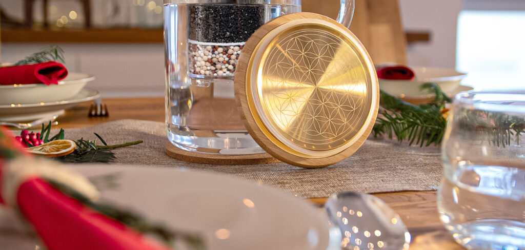 Eine Glas-Wasserfilterkanne mit goldenem Deckel und Lebensblume-Gravur steht auf einem festlich geschmückten Weihnachtstisch.