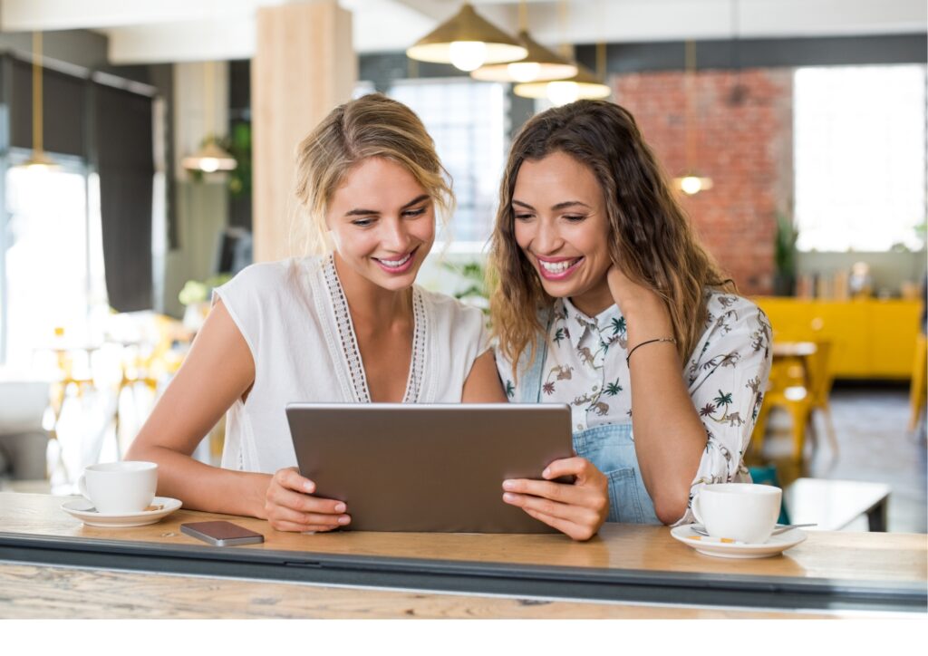 Zwei Freundinnen sitzen lächelnd in einem Café und entdecken gemeinsam auf einem Tablet das Lotus Vita Empfehlungsprogramm.