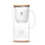 Eine elegante Glas-Filterkaraffe mit Bambusdeckel und Kork-Untersetzer, die das Lotus Vita Logo und das Symbol der Blume des Lebens zeigt.